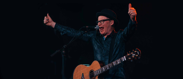 Neil Brophy performing live on stage with an acoustic guitar and engaging the audience 